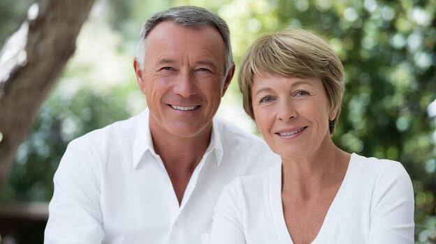 Smiling couple in white shirts outdoors under green trees.